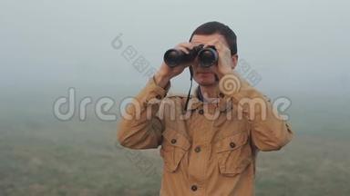 一个身穿卡其色外套的年轻红发男子，手持望远镜，看日出和日落。 四周都是雾